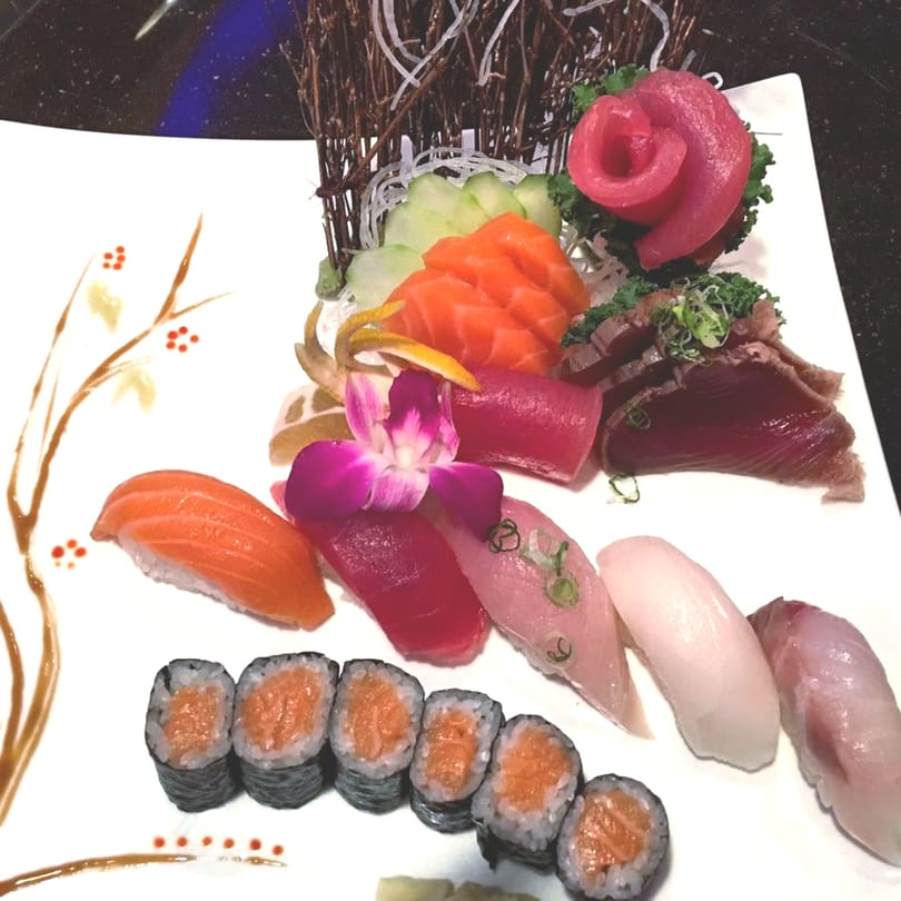 A white rectangular plate holds an assortment of sushi, sashimi, and maki rolls, garnished with an orchid.